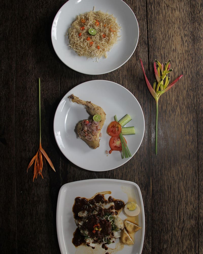 Three Balinese dishes, bulung, ayam betutu and tipat cantok, on a wooden table with heliconia flowers