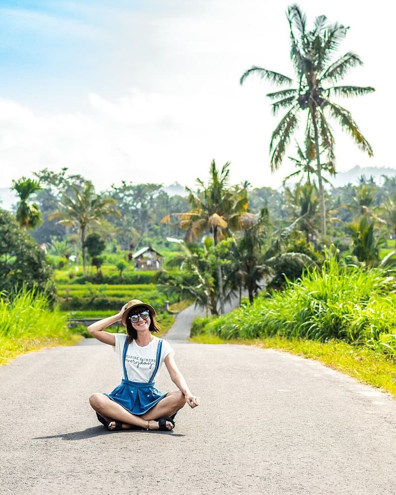 Woman in a straw hat sitting on a quiet palm-lined road between rice fields in Bali